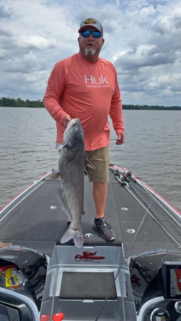 An angler stands atop a boat in the Suwannee River holding a large catfish.